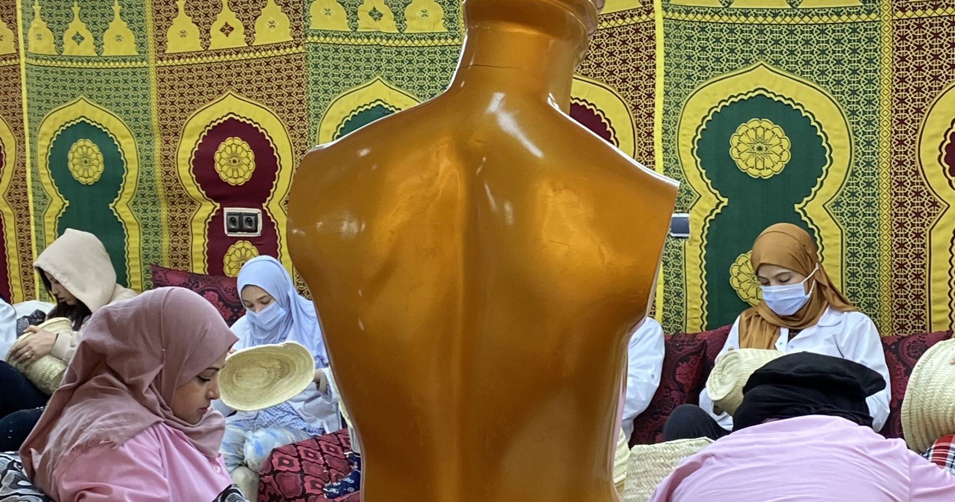 Salle d'atelier tendue de tapisseries à motifs, un mannequin doré au premier plan, des artisanes au travail