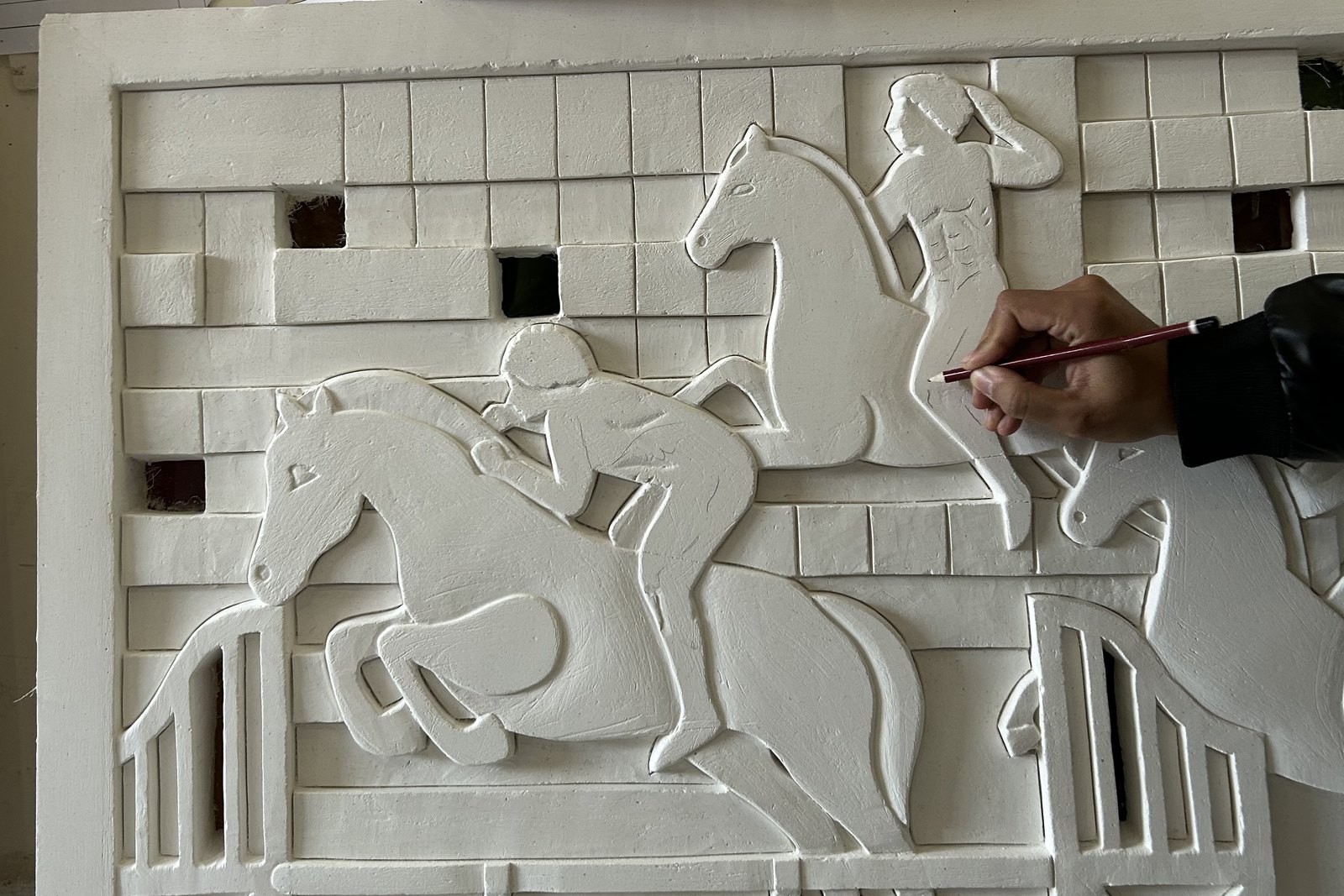 Main de l'artiste dessinant au crayon sur un bas-relief de plâtre représentant des cavaliers à l'obstacle