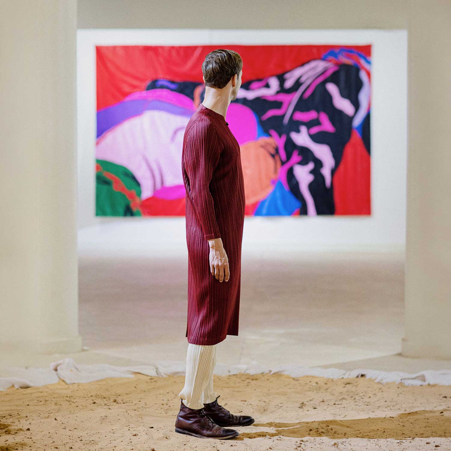 Un visiteur debout, de profil, devant une grande tenture rouge et rose accrochée au mur d'une salle au sol de sable