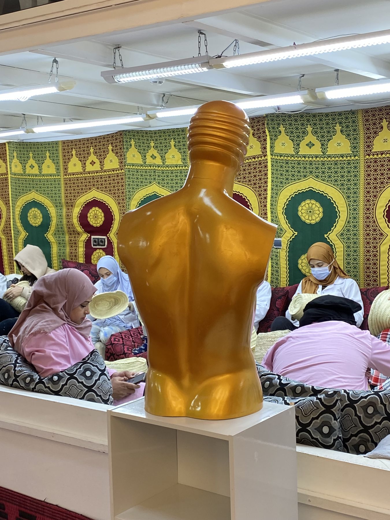 Salle d'atelier tendue de tapisseries à motifs, un mannequin doré au premier plan, des artisanes au travail