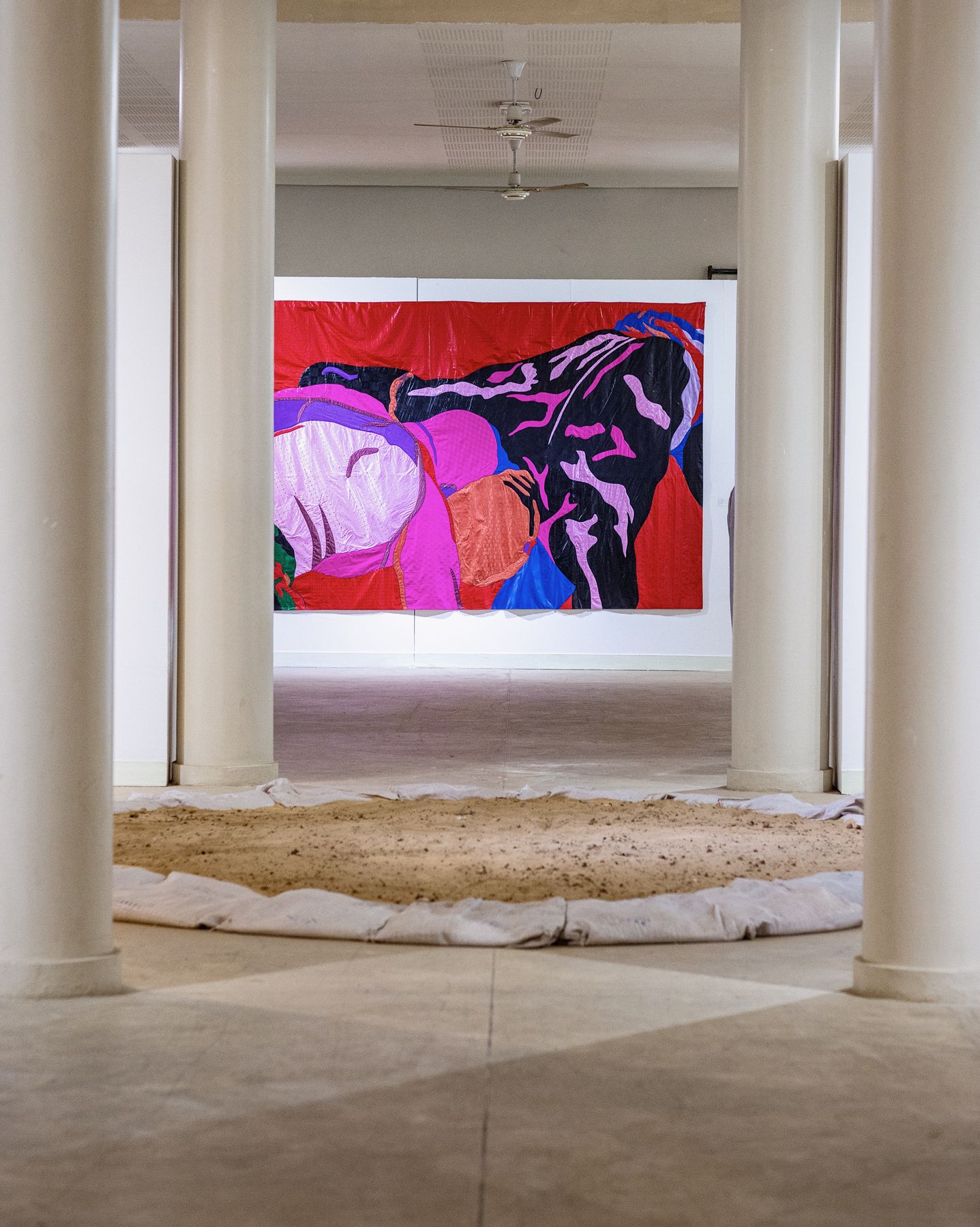 Vue d'exposition : une grande tenture aux figures roses et bleues au fond d'une enfilade de colonnes, sol de sable