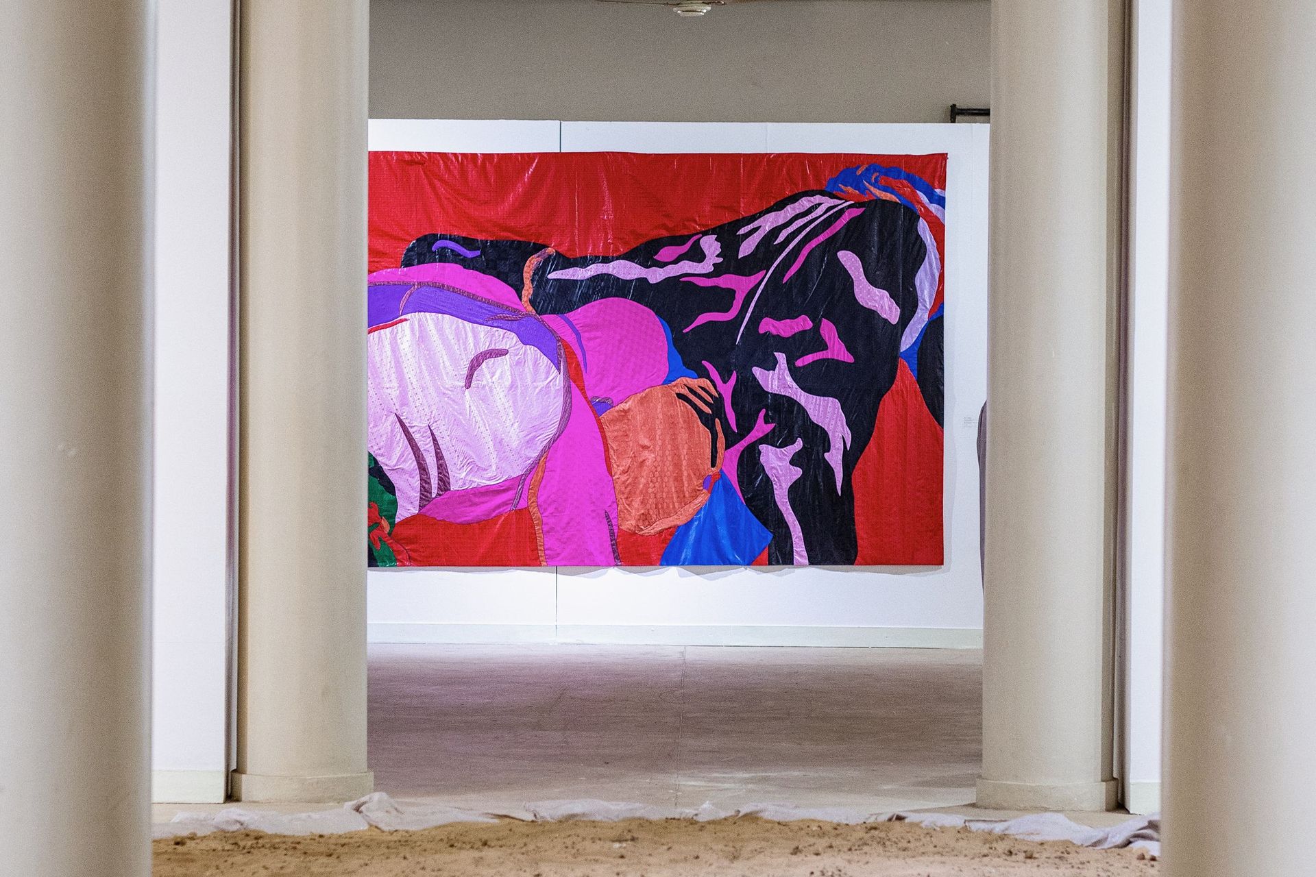 Grande œuvre textile accrochée entre les colonnes d'une salle d'exposition, sol de sable