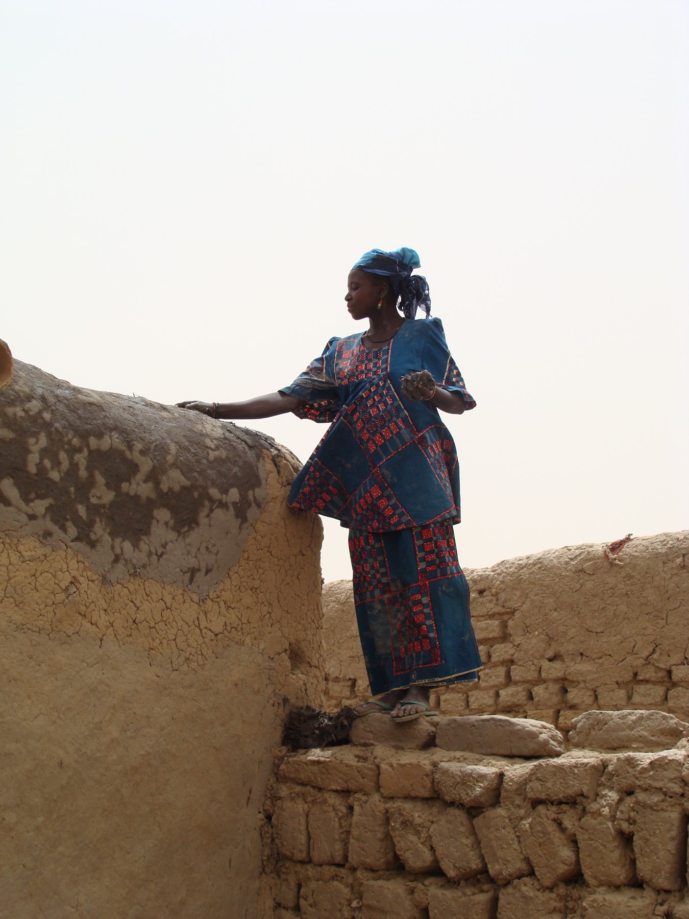 Photographie d'Amélie Essessé : une femme en robe bleue à motifs, debout sur un mur de briques de terre, lisse de la main un toit de banco, île de Koutougou, Niger